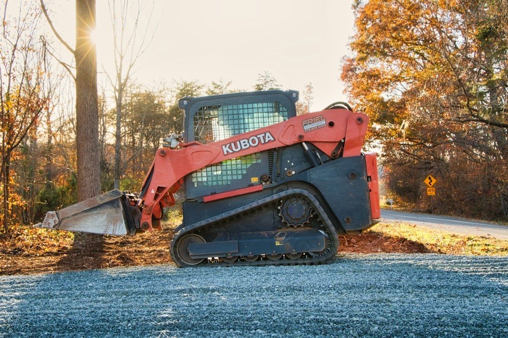 A compact track loader parked on a freshly cleared gravel drive beside autumn woods